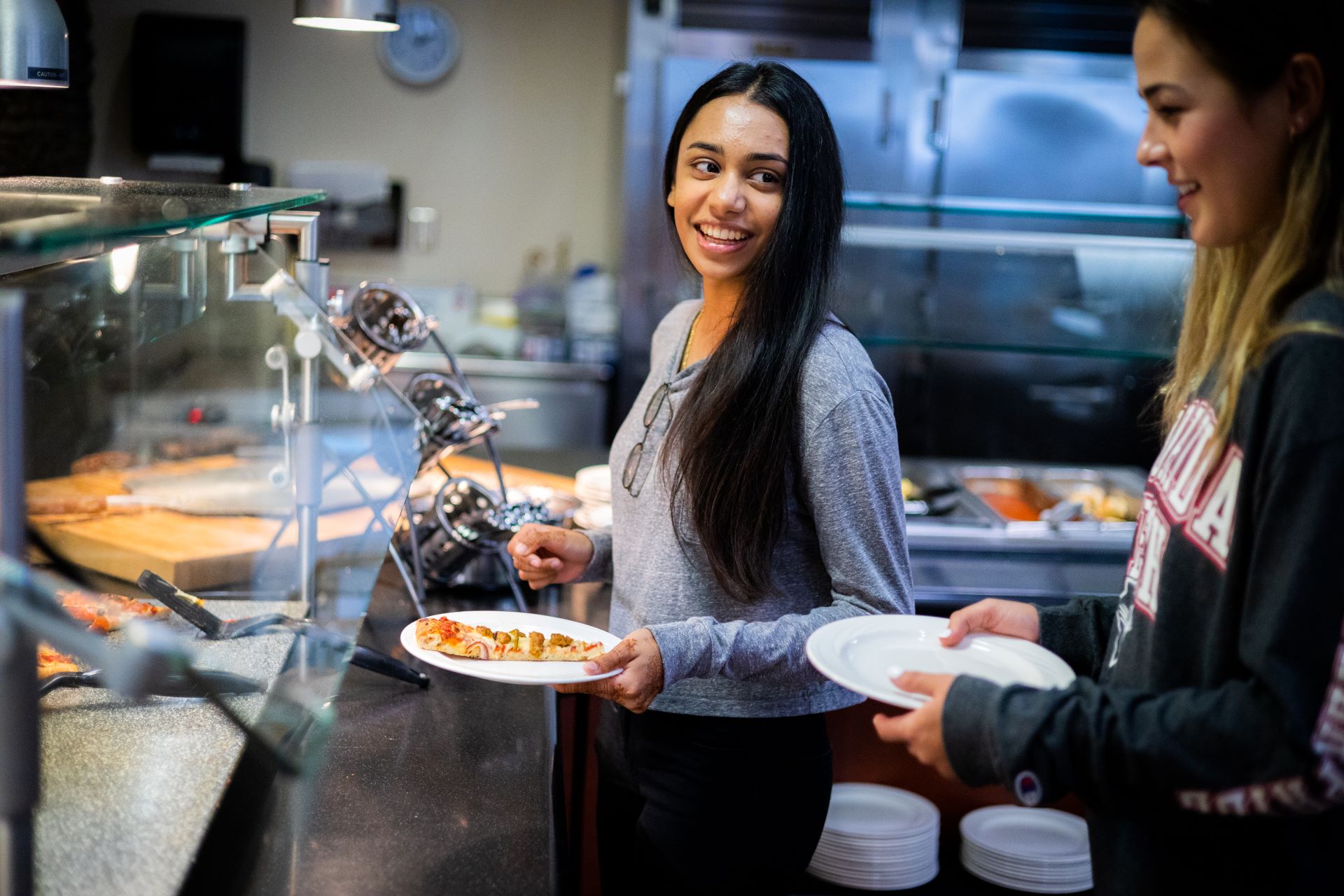 Two young women, one smiling at the camera, serving themselves food at a college dining hall buffet
